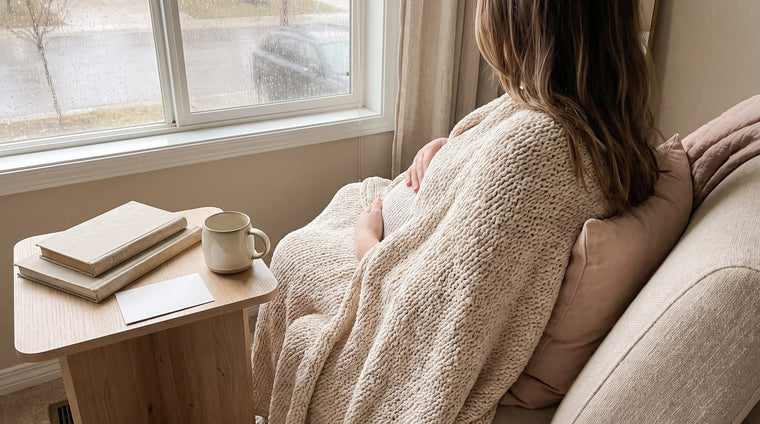 Schwangere mit Decke am regnerischen Fenster, eine Hand am Bauch, gedämpftes Licht