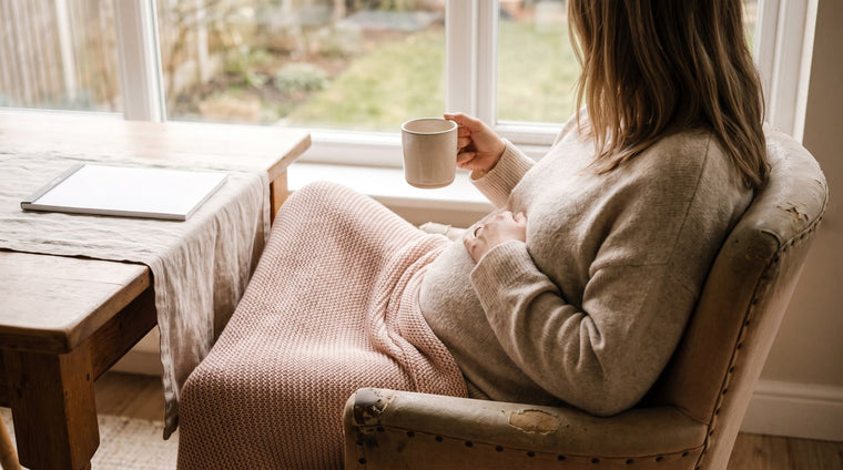 Schwangere sitzt entspannt mit Teetasse am hellen Fenster, eine Hand auf dem Bauch, weiche Decke