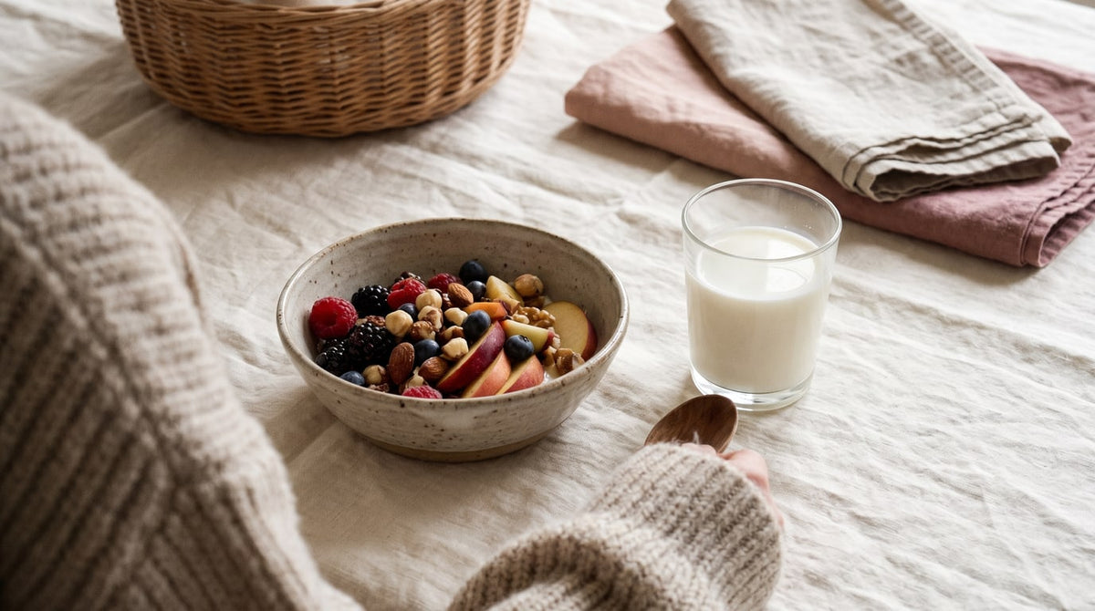 Schale mit frischem Obst und Nüssen neben einem Glas Milch auf cremefarbenem Leinen im warmen Licht