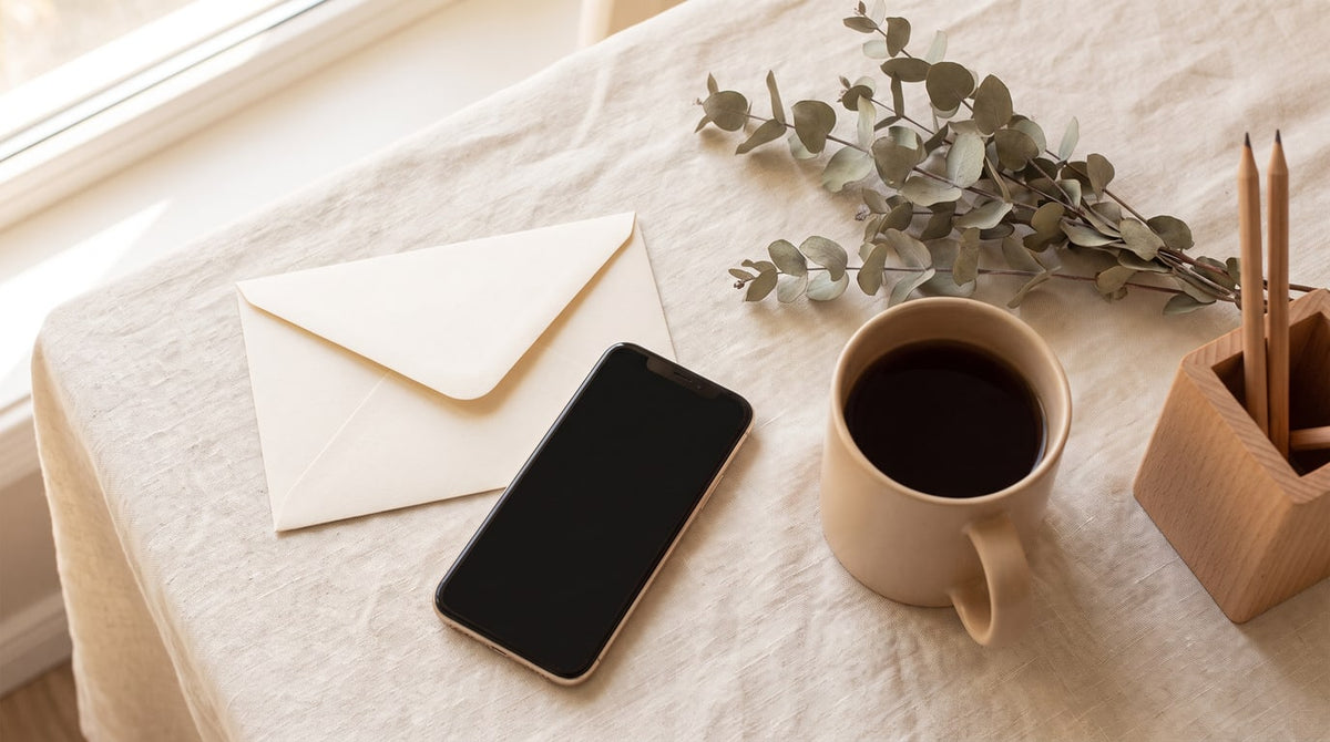 Cremefarbener Briefumschlag, ein Smartphone mit dunklem Display, Teetasse und Eukalyptus auf Leinen im warmen Tageslicht