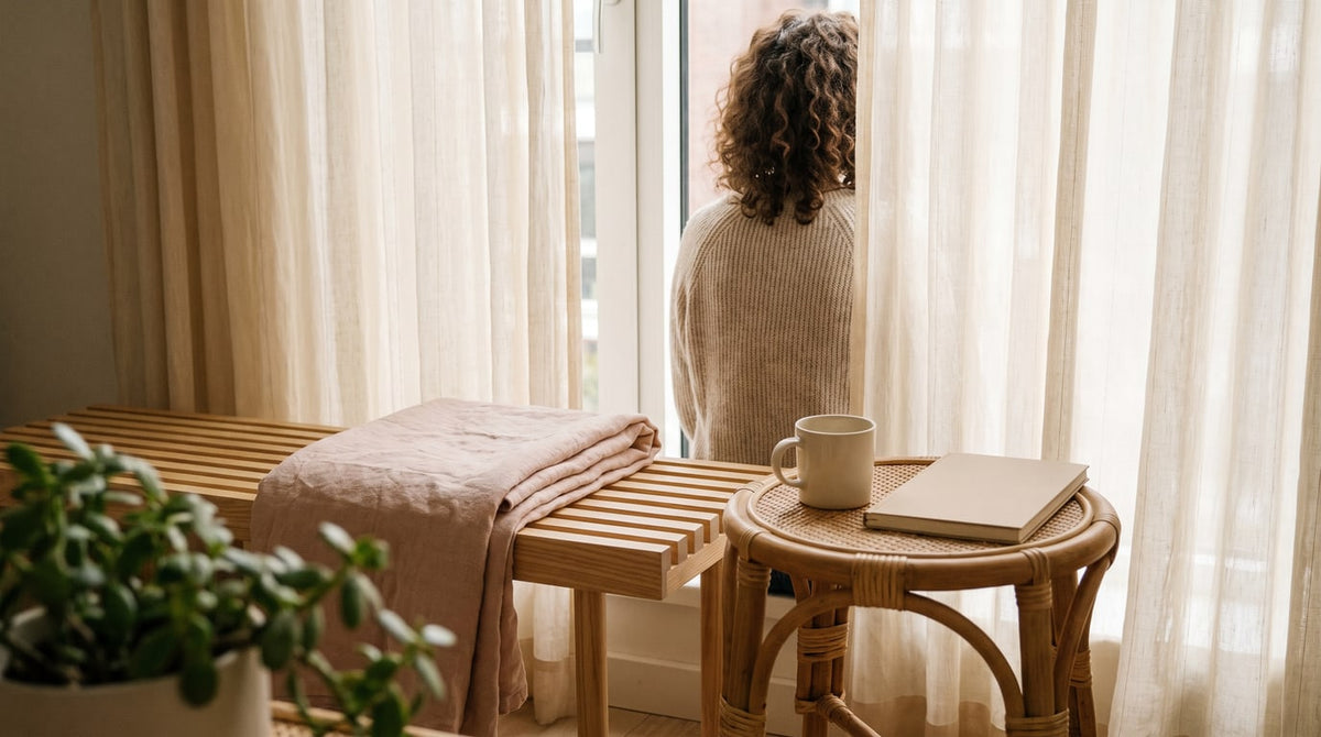 Heller Wartebereich mit Holzbank, blassrosa Decke, cremefarbener Tasse und Notizbuch am Fenster